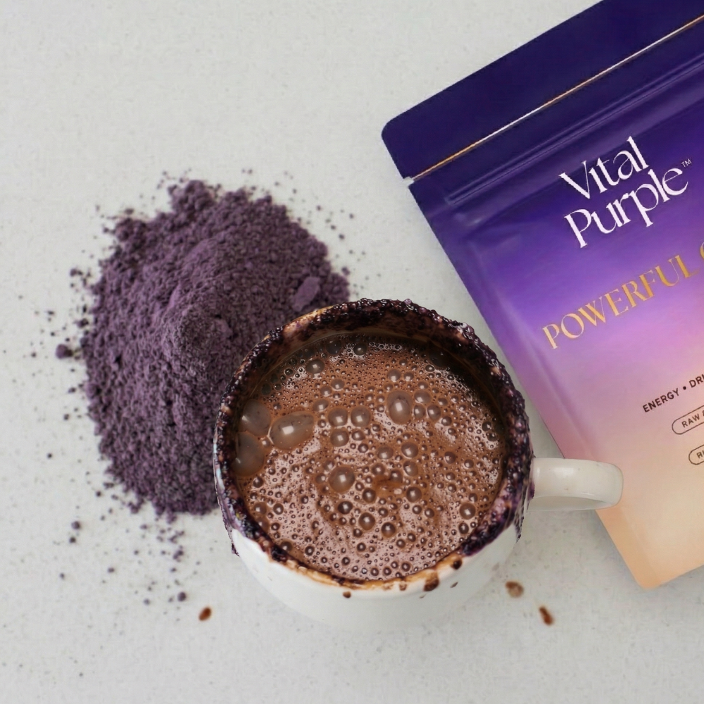 A cup of frothy dark brown beverage with a white handle, next to a purple pouch labeled Vital Purple and a pile of loose purple powder on a light surface.