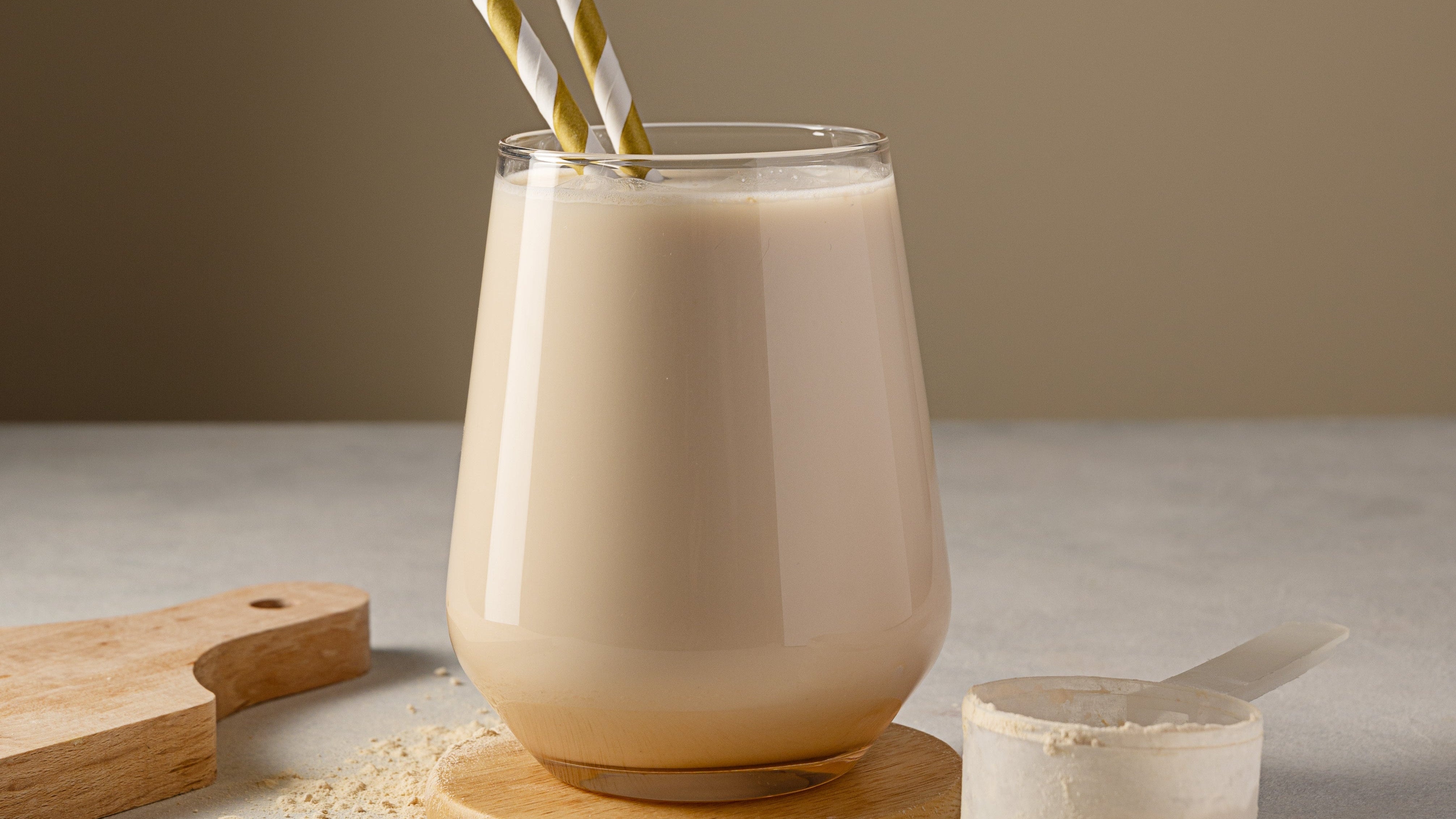 Glass of creamy beverage with straws on a wooden coaster, surrounded by a wooden cutting board and container of powder.